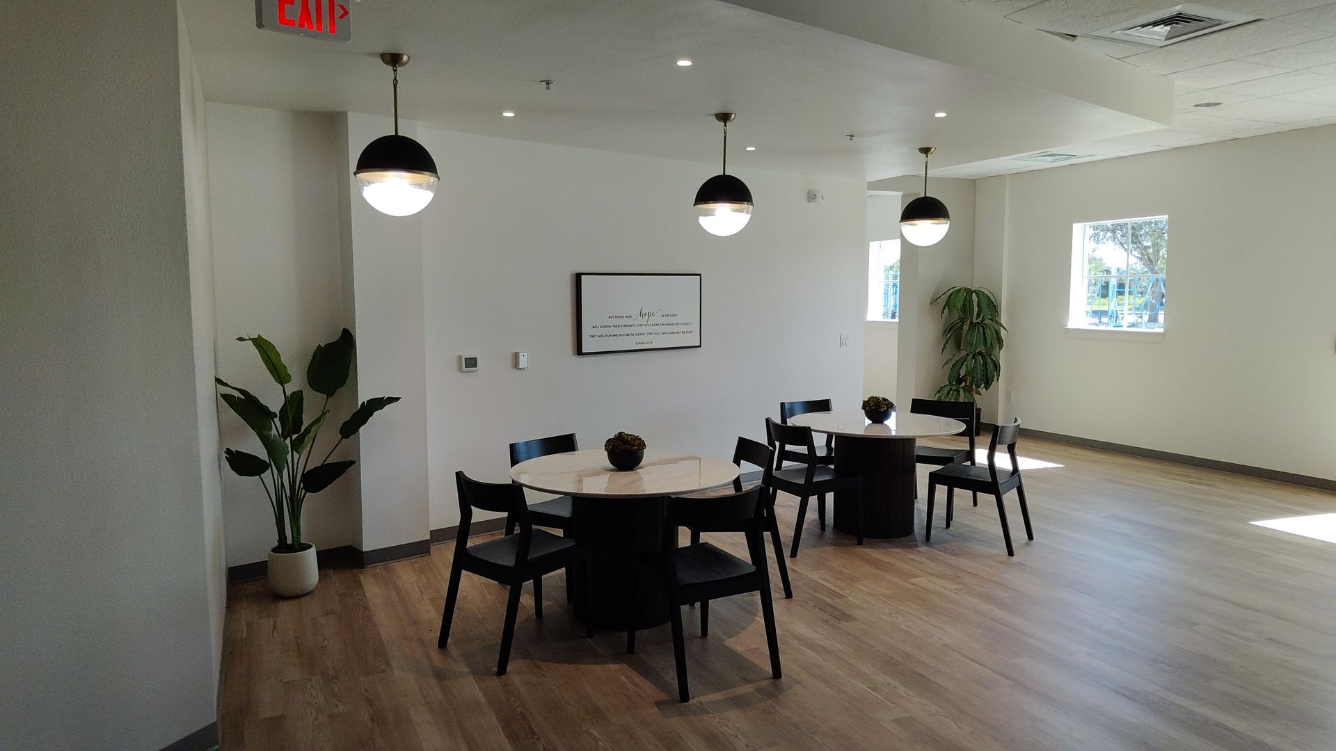 Modern office lounge with round tables, black chairs, pendant lights, plants, and wood floors.