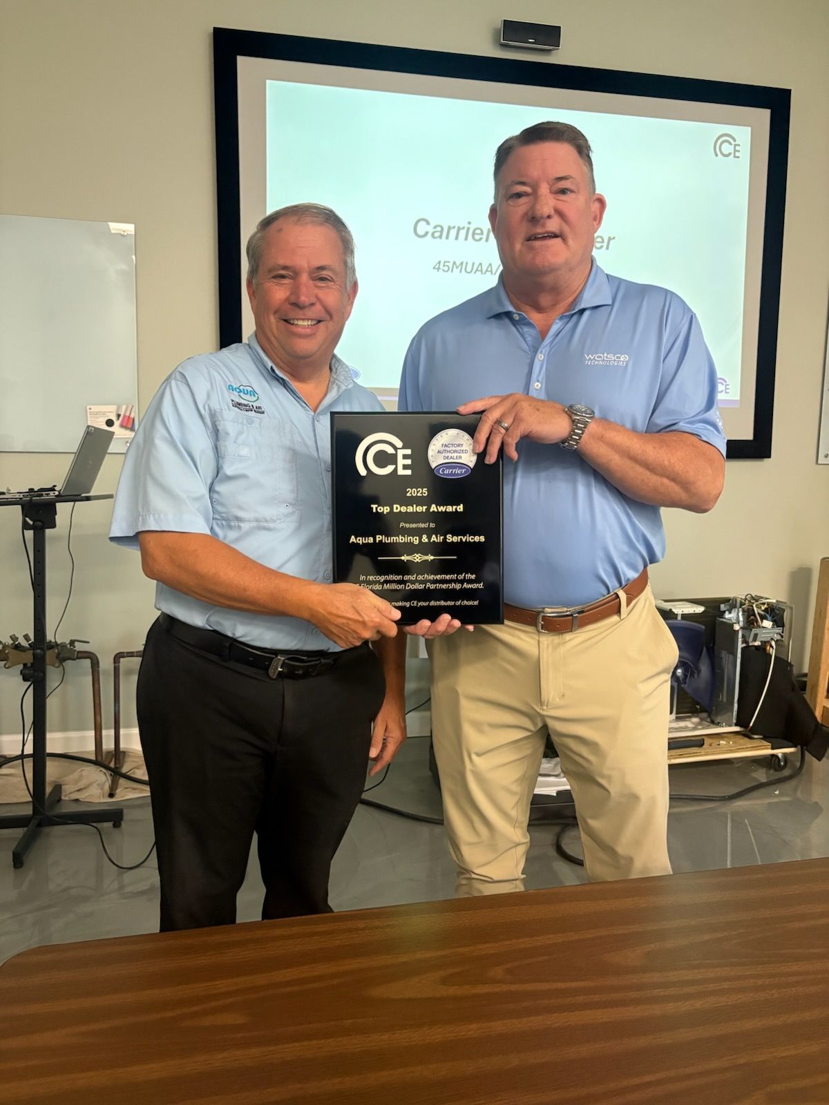 Two men hold a plaque in front of a projector screen in a meeting room.