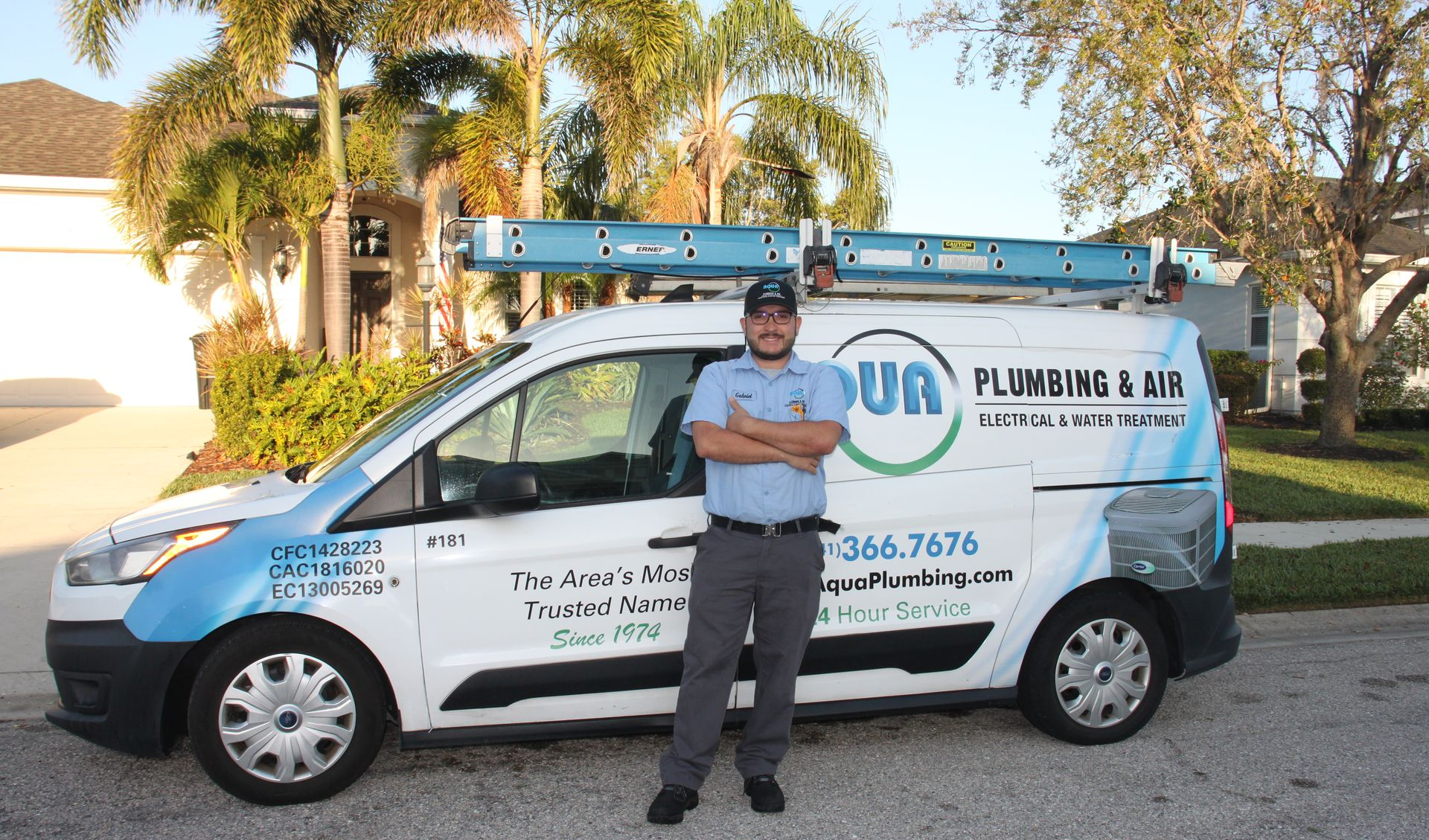 Plumber standing beside a white service van with blue lettering and a ladder on top, parked outdoors.