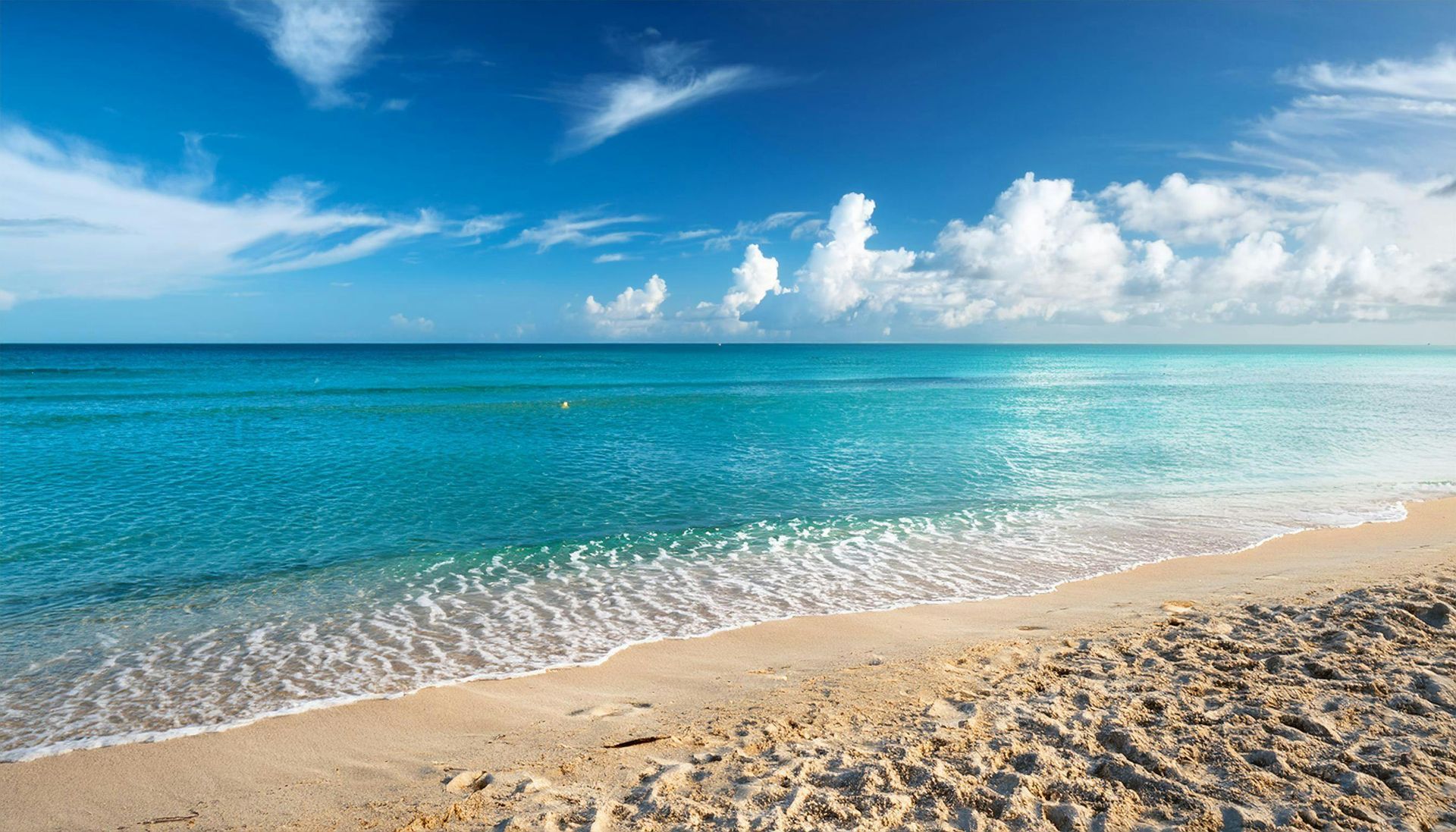 A bright, sunny beach with calm, turquoise water meeting white sand under a sky filled with scattered white clouds.