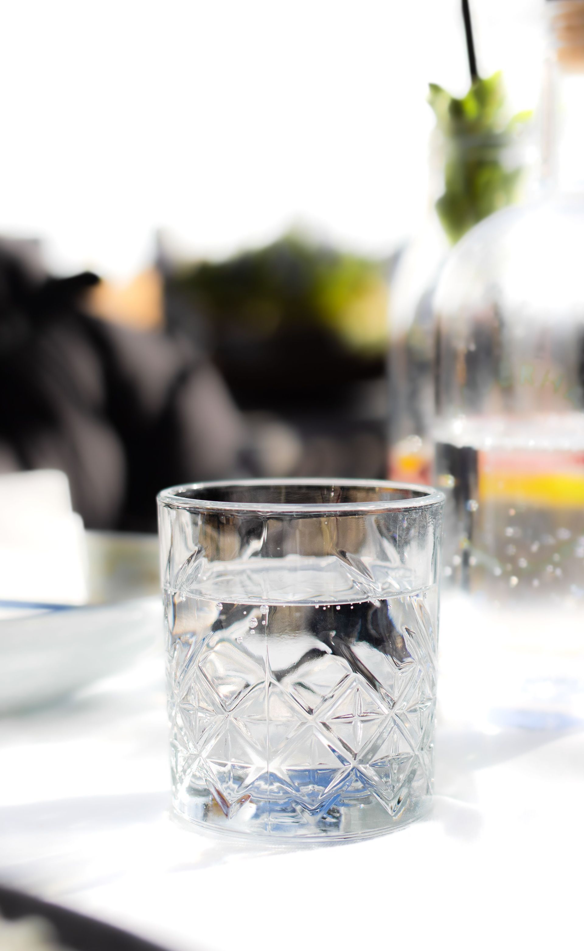 Clear glass tumbler on a sunlit table with a blurred vase and greenery in the background