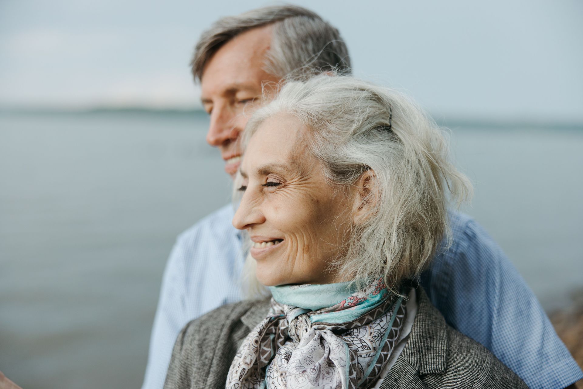 Two people standing by the water, one smiling in a scarf and sweater, with a man behind her looking out over the sea