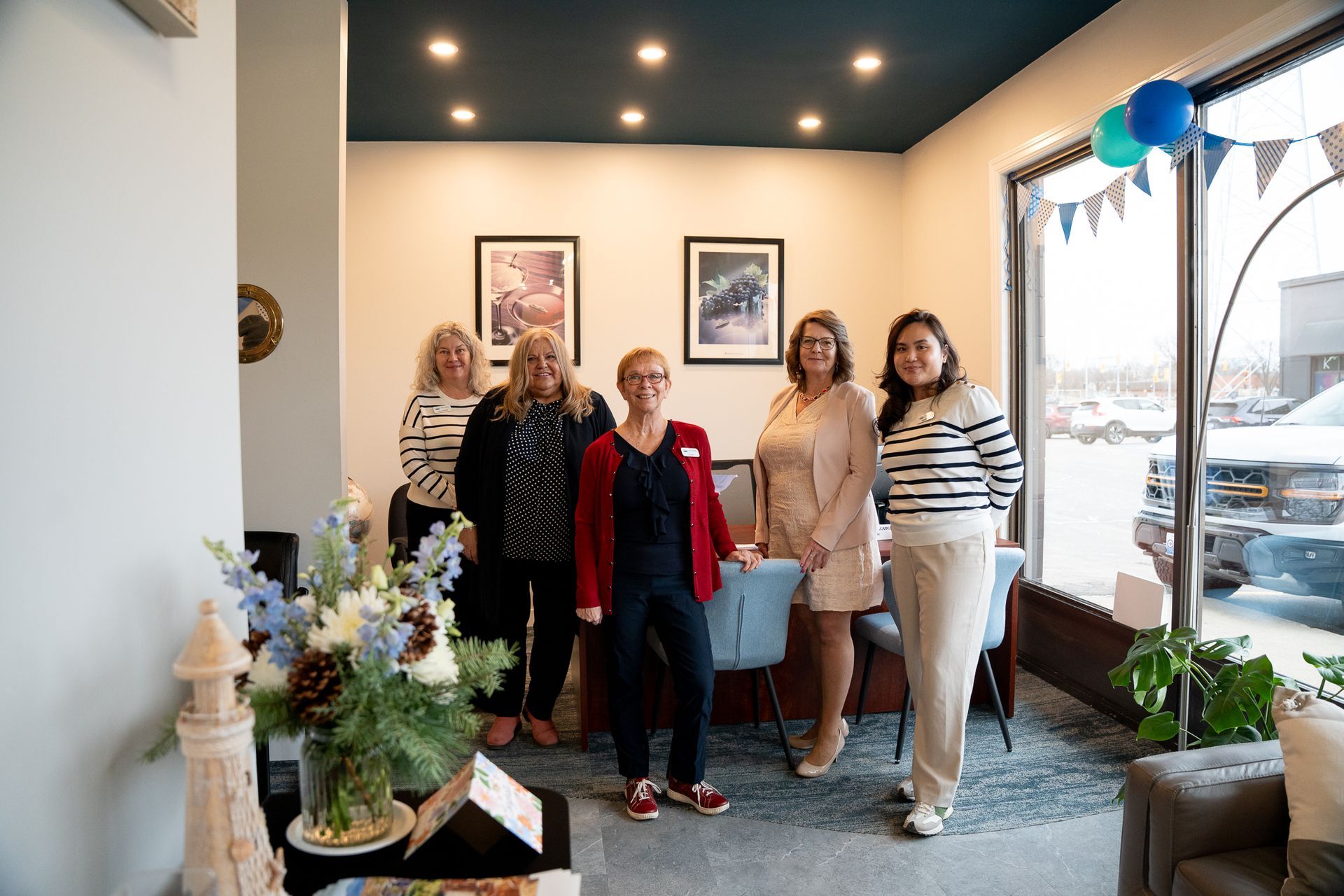 Five people standing in a bright room with framed art, a table, and flowers near a window with bunting.