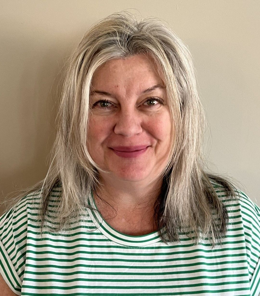 Smiling woman with wavy blonde hair wearing a green-and-white striped shirt against a beige wall