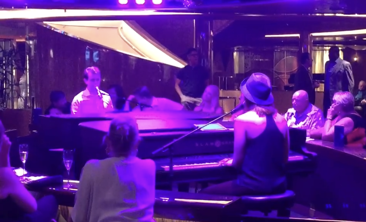 singing next to a piano A woman is singing into a microphone in front of a piano in a dark room.