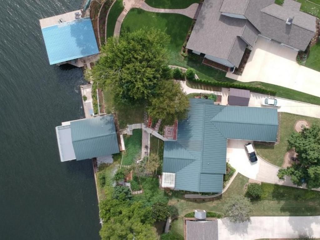 An aerial view of a lakefront property featuring a house with a green roof, a dock with a blue roof, and a gravel driveway.