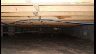 A crawl space beneath a wooden floor structure, showing floor joists, wiring, and a dark dirt ground.