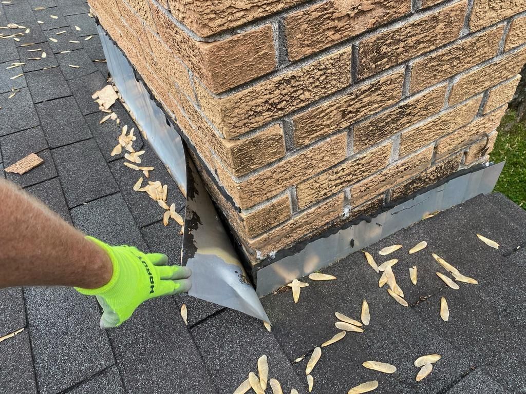 A gloved hand pulls back loose metal flashing on a residential chimney where it meets the shingled roof.