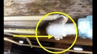 A yellow circle highlights a metal elbow pipe fitting leaking a single drop of liquid in a basement crawl space.