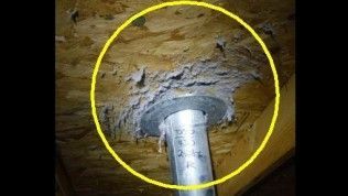 A silver metal pipe passing through a wooden attic ceiling with grey insulation fibers clustered around the opening.