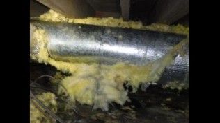 A metal HVAC duct in a dark attic space surrounded by loose, yellow fiberglass insulation.