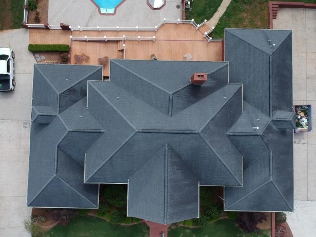 An aerial view of a dark grey shingled residential roof with a chimney and a small portion of a pool deck visible.