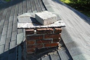 A small, square brick chimney on a shingled roof, topped with a stone slab and a single heavy concrete block.