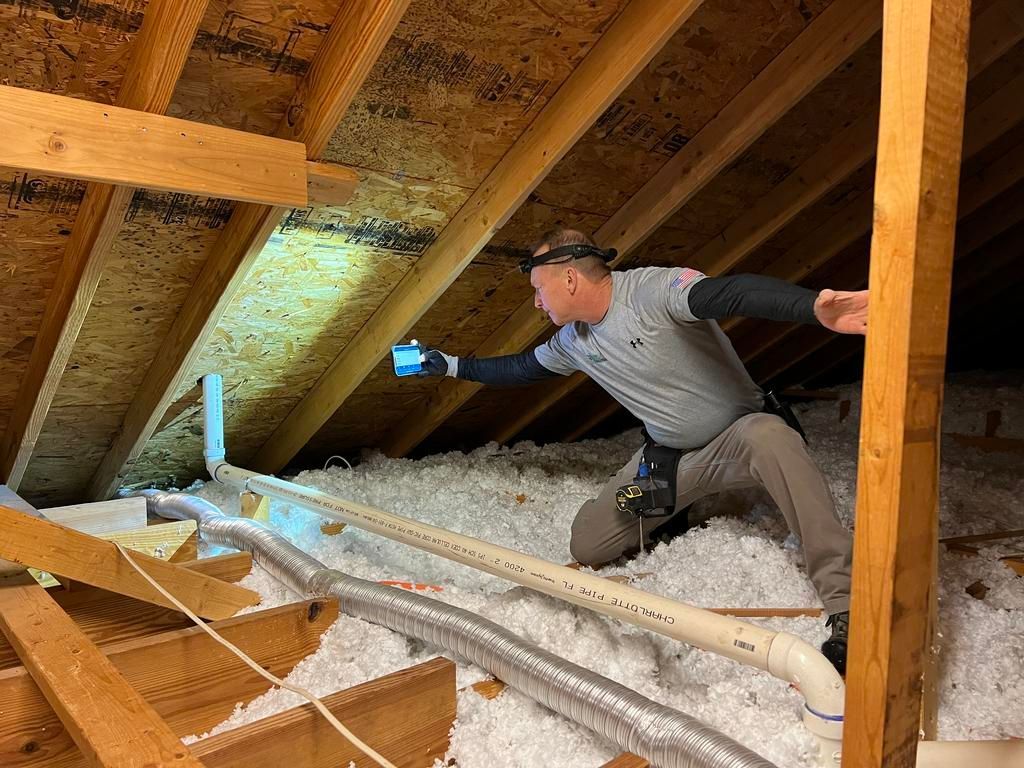 An inspector kneeling in an attic uses a device to check for moisture on wooden beams and sheathing near a vent pipe.