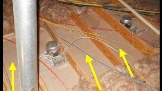 A high-angle view of attic joists and wiring with three yellow arrows pointing to electrical cables running between beams.