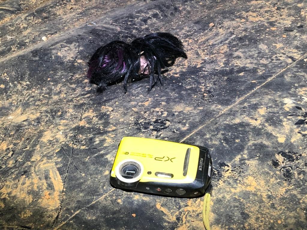 A large, black spider resting on a dark, textured surface next to a small, yellow digital camera.
