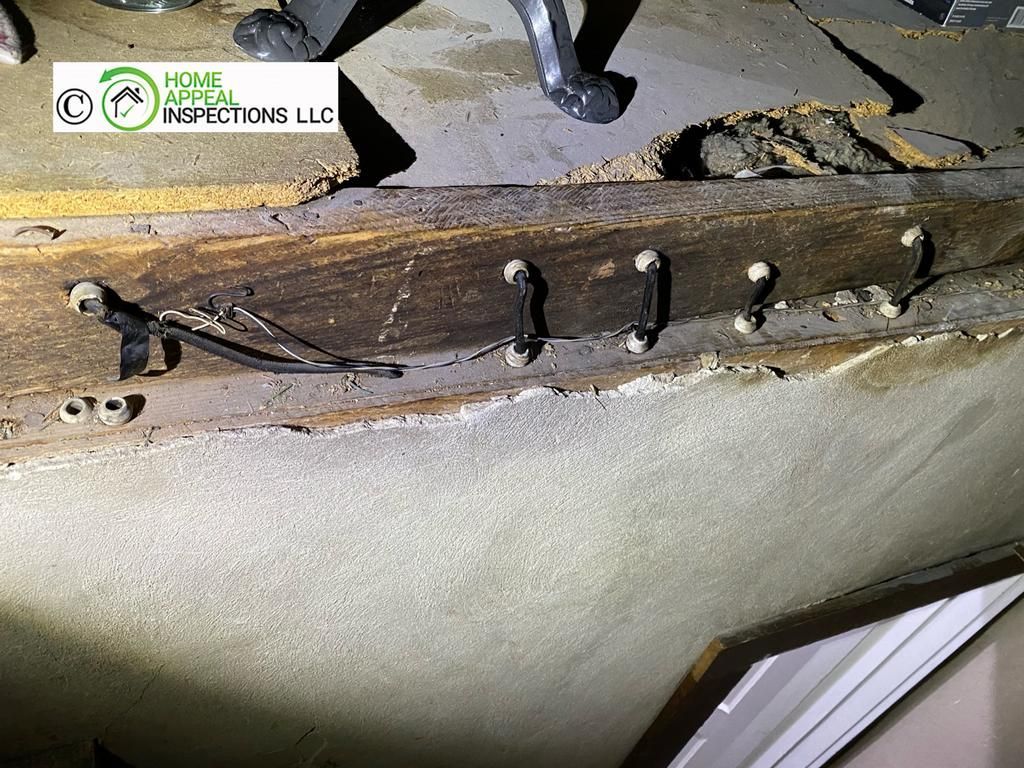 Black electrical wiring stapled in a series of loops across a wooden beam against a concrete foundation.