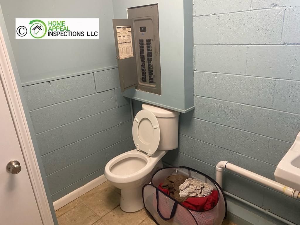 Electrical panel mounted on a blue wall directly above a toilet in a small room with a laundry hamper.