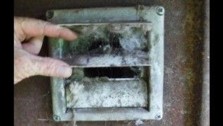 A person's hand opens a wall dryer vent cover that is clogged with a thick buildup of gray lint.