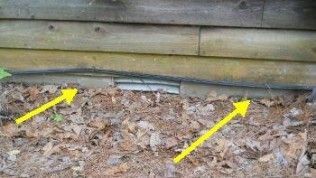 A close-up of a cabin's foundation with yellow arrows pointing to a horizontal gap and vent beneath the wooden siding.