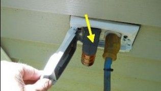 A hand opens a protective outdoor electrical box cover, showing a power plug and a copper-colored connector marked by a yellow arrow.