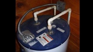 An electric water heater with white PVC piping connections on top, situated in a room with wood-paneled walls.