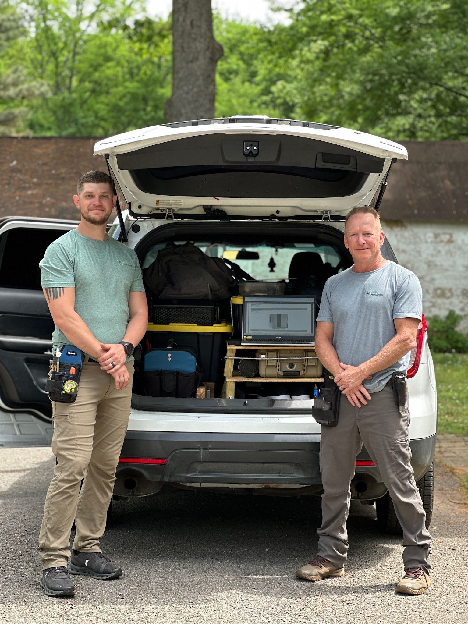 Two men standing behind an open SUV trunk filled with gear and a laptop setup outdoors.
