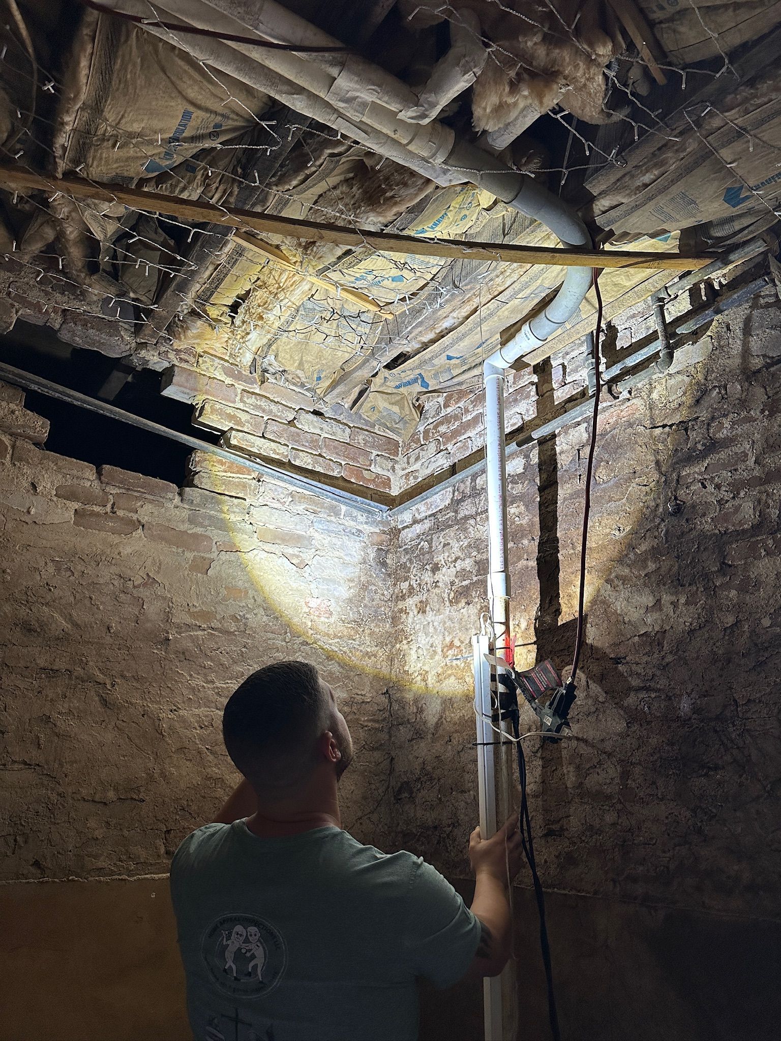 Worker inspecting damaged ceiling with flashlight in a dim attic, exposed beams and hanging cables.