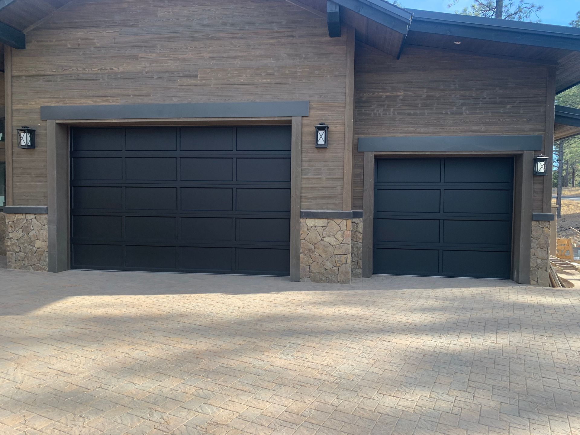 A house exterior with two black horizontal-slatted garage doors set in a wood-paneled building with stone corner accents.