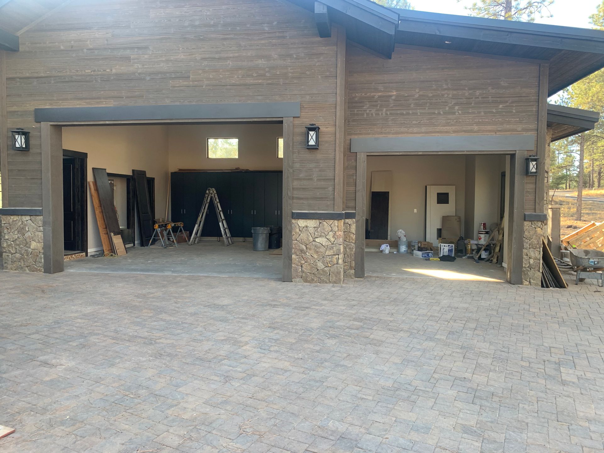 A rustic garage with wood siding and stone pillars, featuring two open bays with construction equipment visible inside.