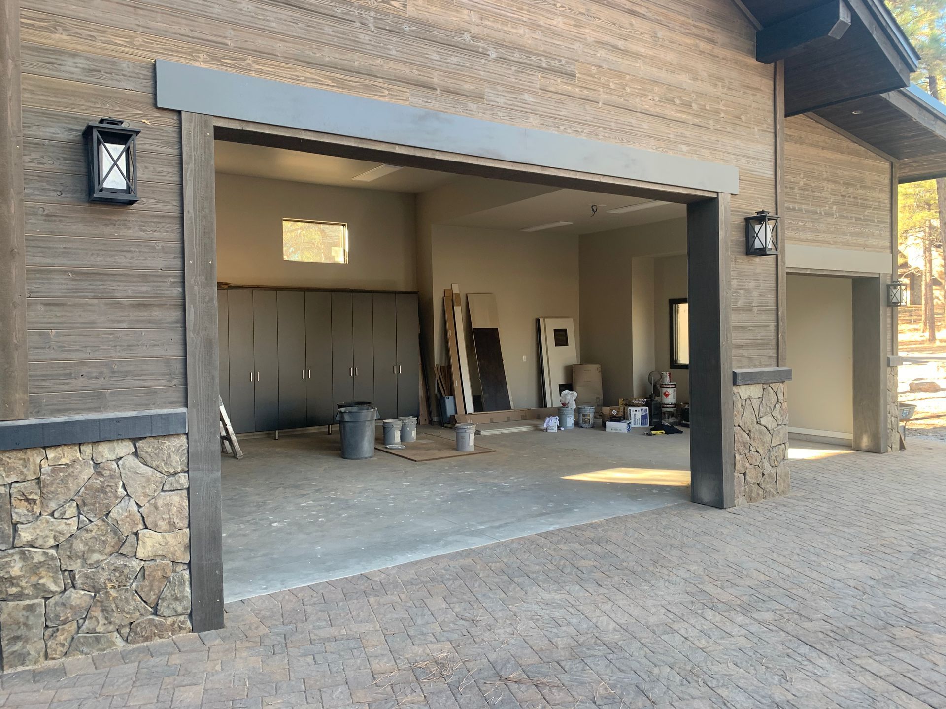 An open two-car garage with stone siding, wood trim, outdoor lanterns, and construction supplies inside.