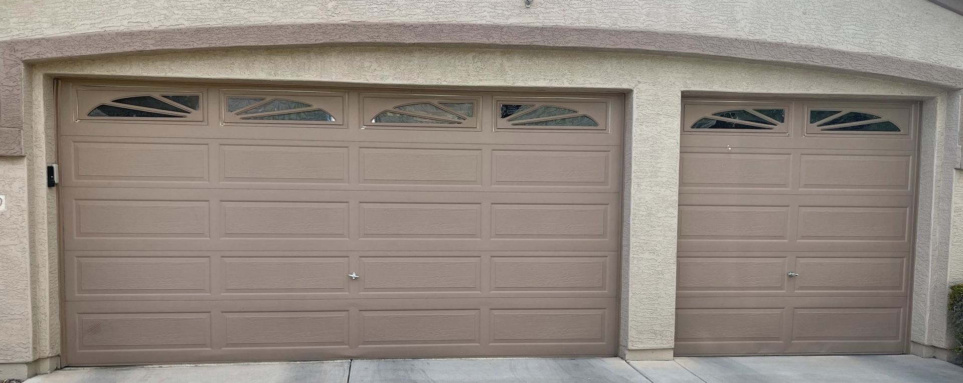 Tan double and single car garage doors with decorative arched window inserts, set in a beige stucco building exterior.