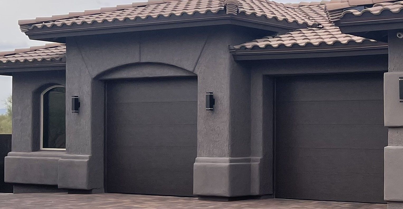A modern house exterior featuring two dark-colored garage doors and a textured gray stucco facade with tile roofing.