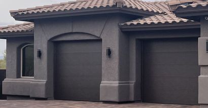 A modern house exterior featuring two dark-colored garage doors and a textured gray stucco facade with tile roofing.