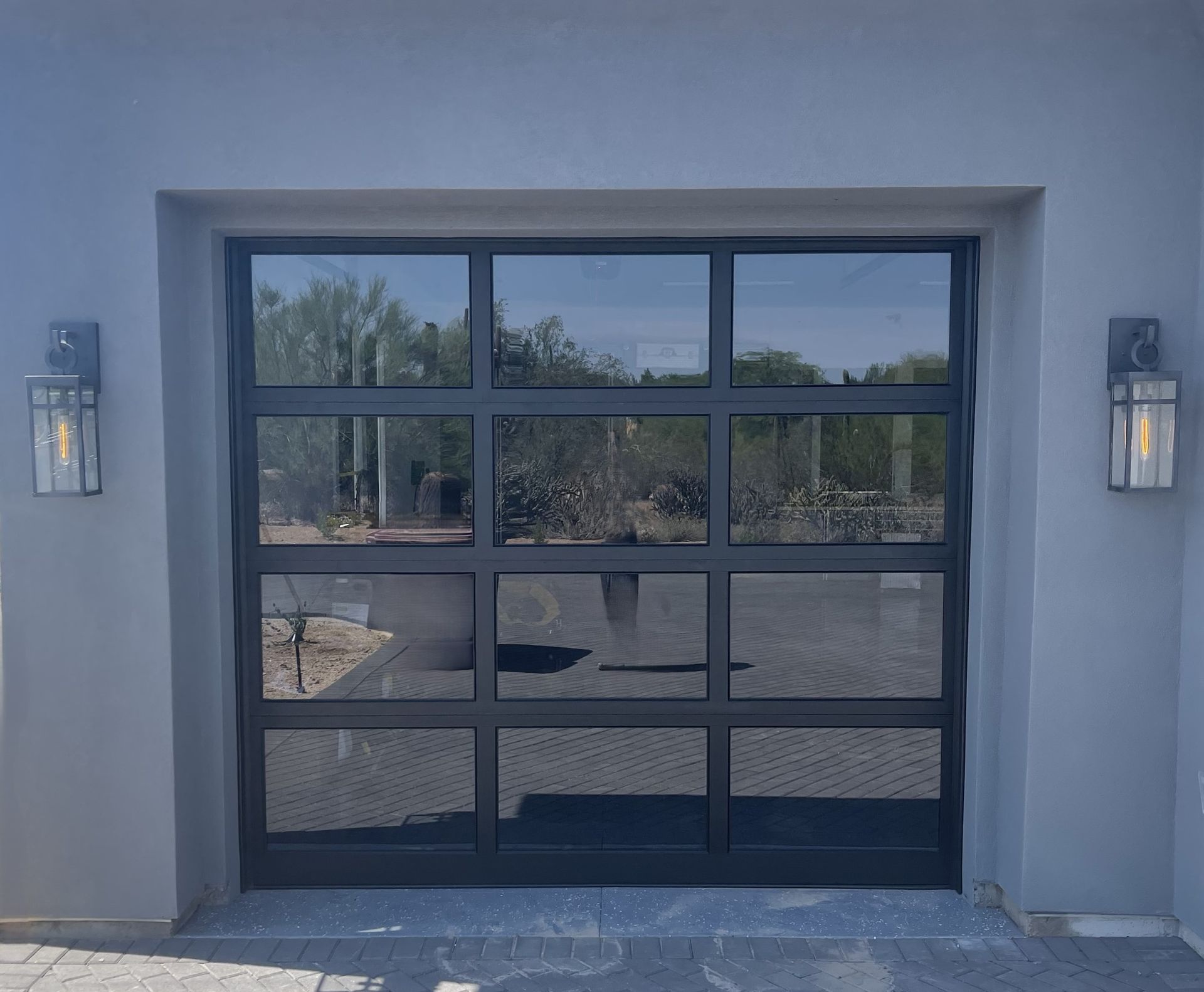 A modern, black-framed garage door with rectangular glass panels, flanked by two outdoor wall lanterns on gray stucco.