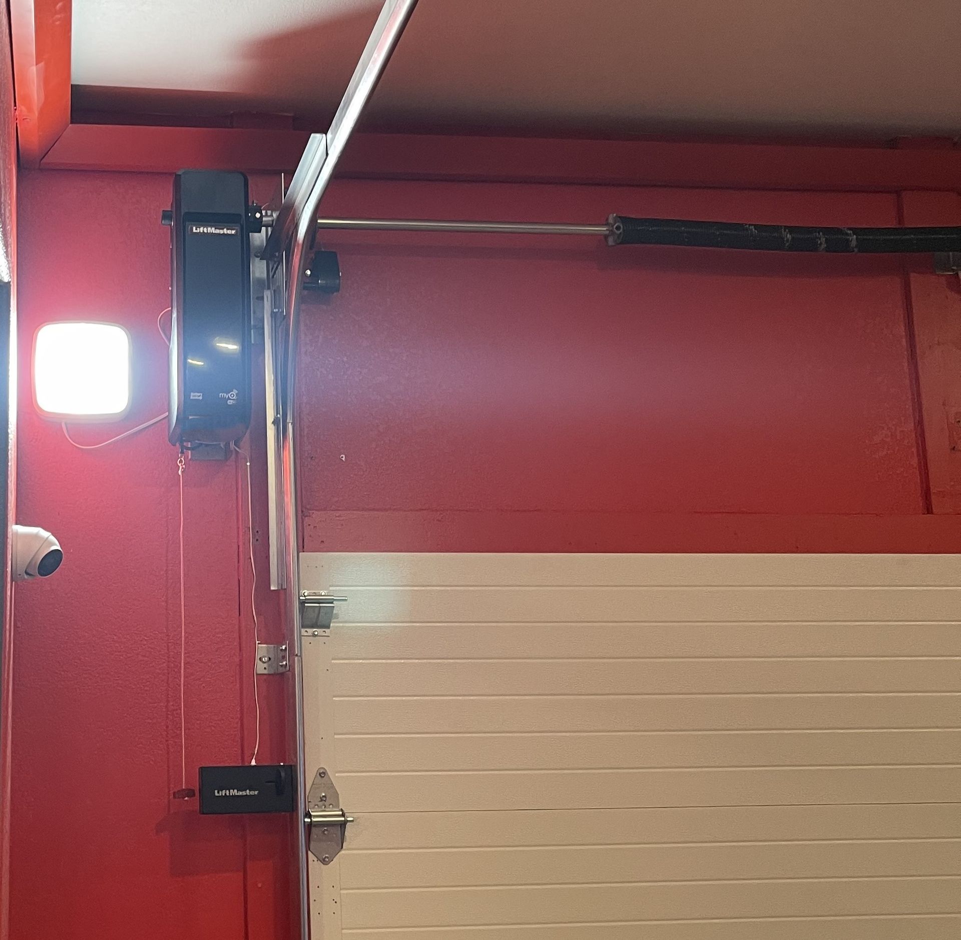 A side-mounted garage door opener and a bright LED light fixture mounted on a red wall above a white garage door.