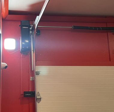 A side-mounted garage door opener and a bright LED light fixture mounted on a red wall above a white garage door.