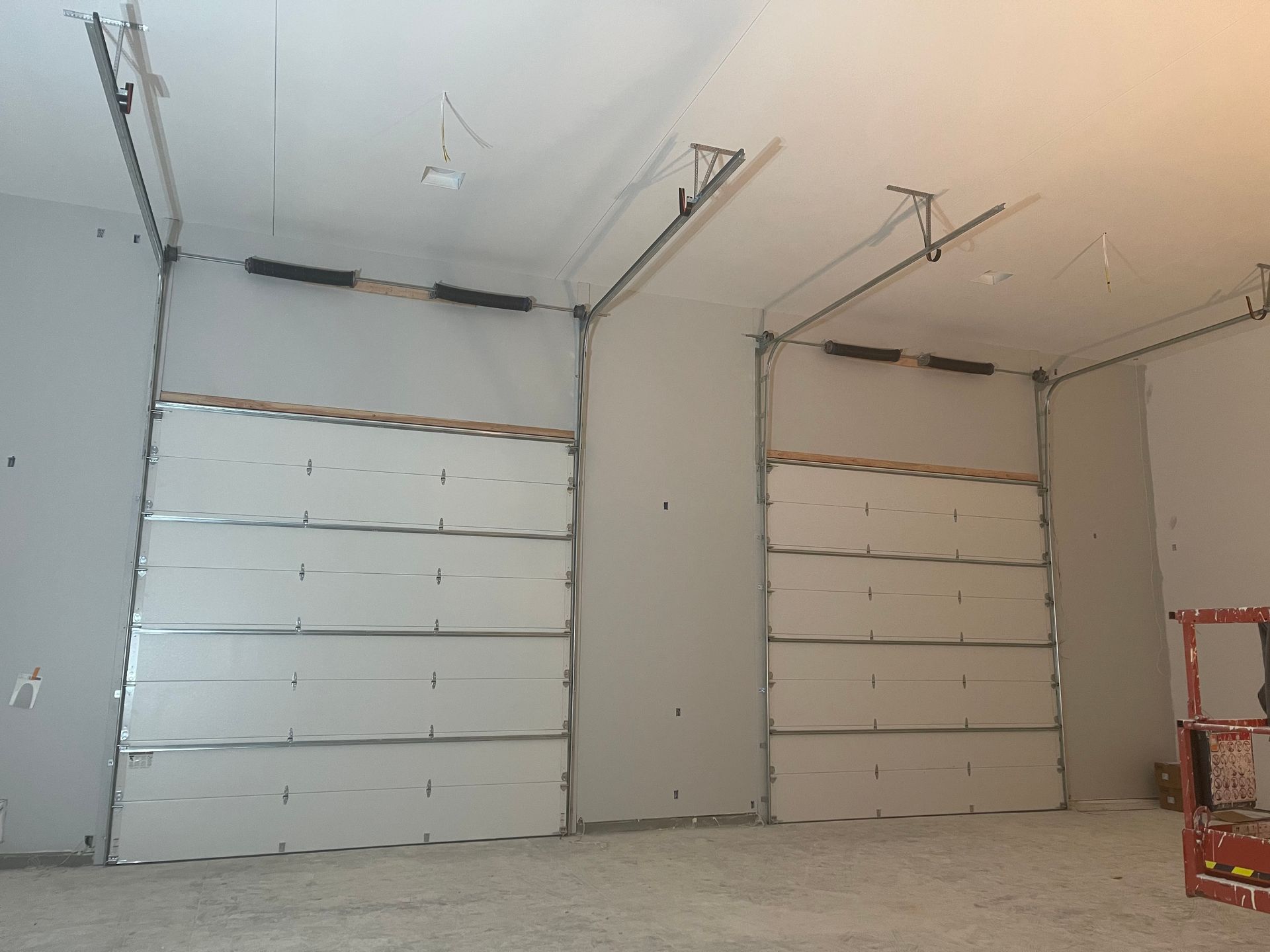 Two white sectional garage doors installed in a room with unfinished drywall and an exposed ceiling structure.