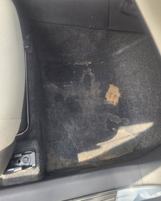 Car floor mat with a worn tan stain and scuffed black carpet near the seat rail