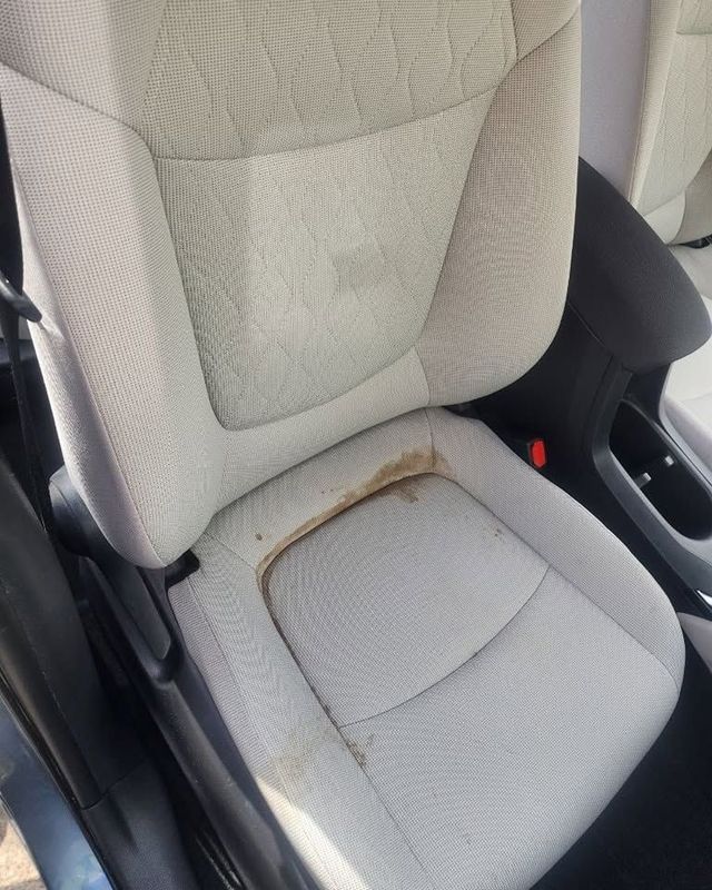 Car seat with a large brown stain on the light gray seat cushion and backrest.