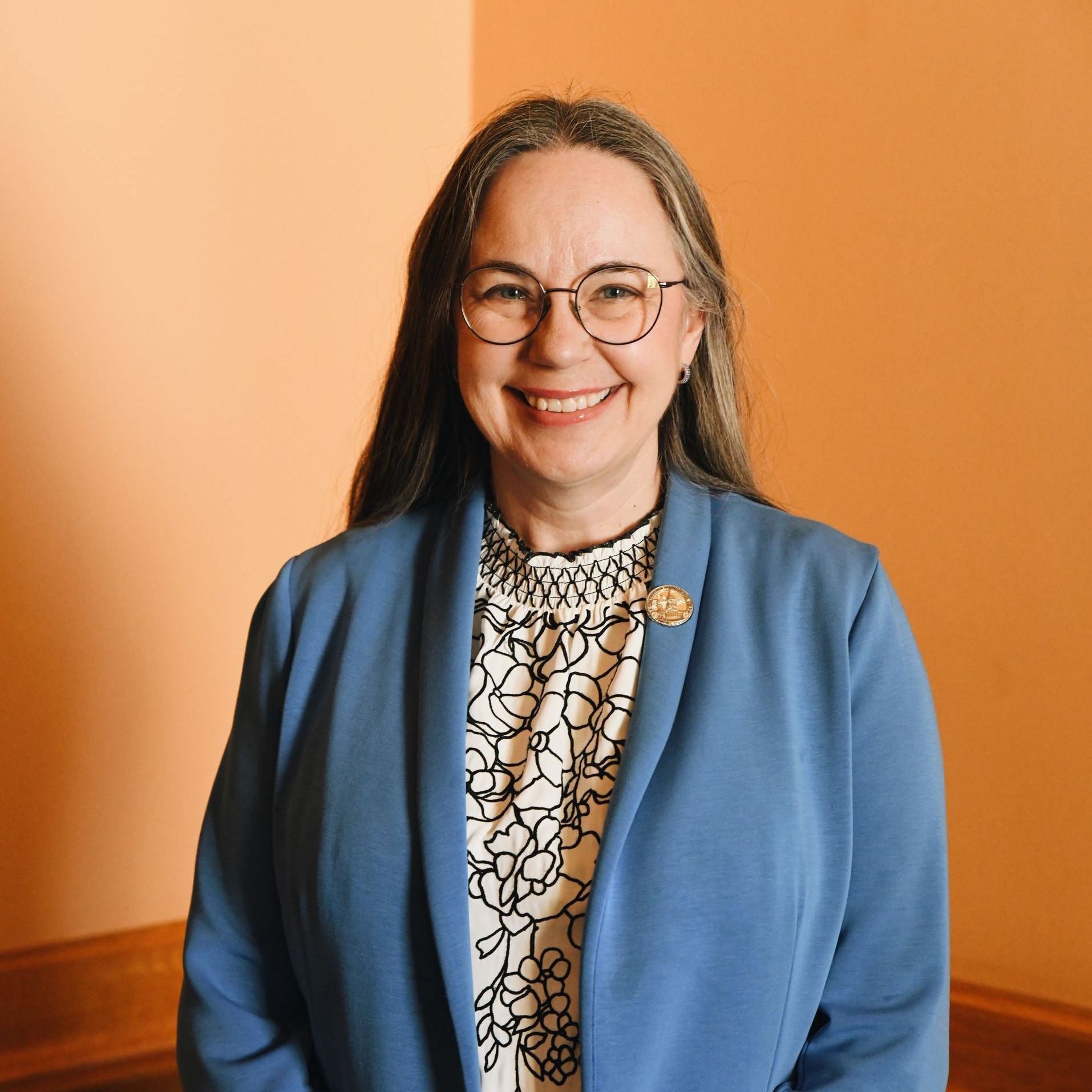 A woman wearing glasses and a blue jacket is smiling for the camera.