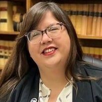 A woman wearing glasses and a black jacket is smiling in front of a bookshelf.