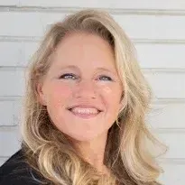 A woman with long blonde hair is smiling in front of a white wall.