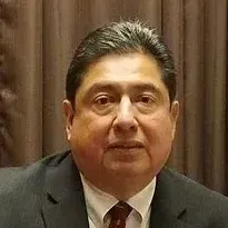 A man in a suit and tie is sitting in front of a brown curtain.