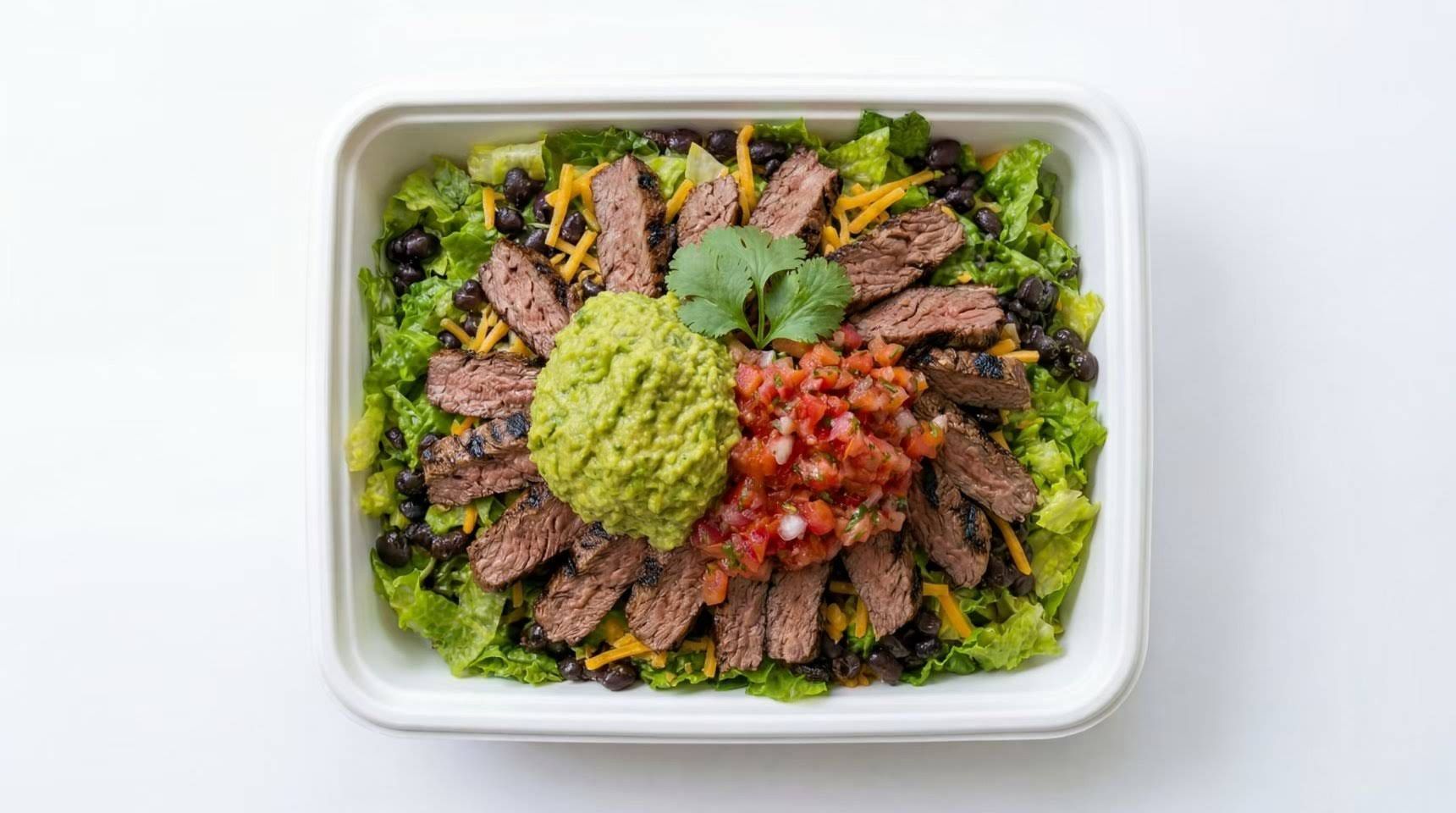White takeout container with chopped steak, lettuce, pico de gallo, and green sauce on a white background