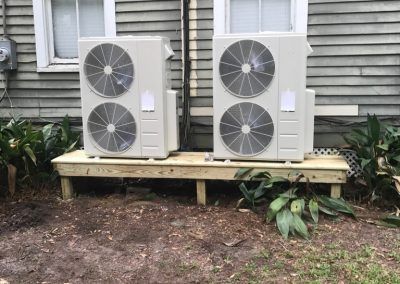 Two air conditioners are sitting on a wooden platform in front of a house.