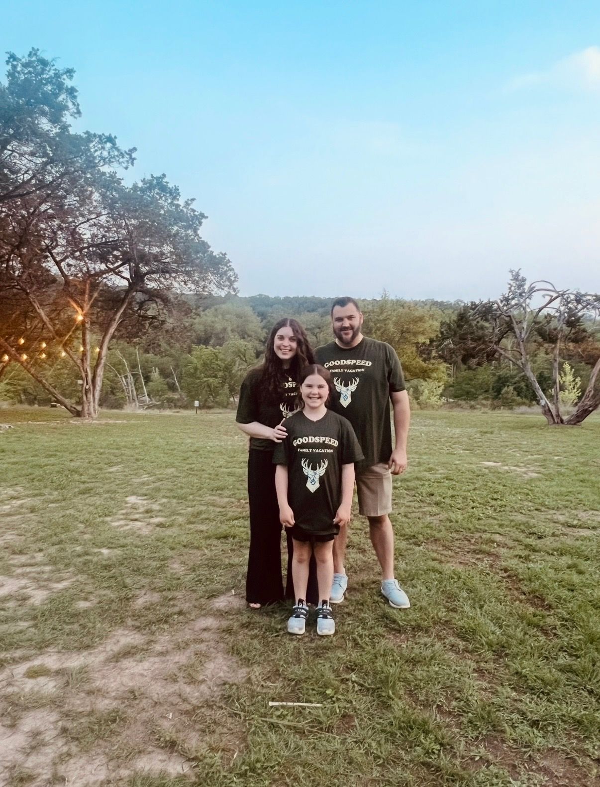 Three people standing on grass at dusk in a park, with trees and soft sunset light behind them