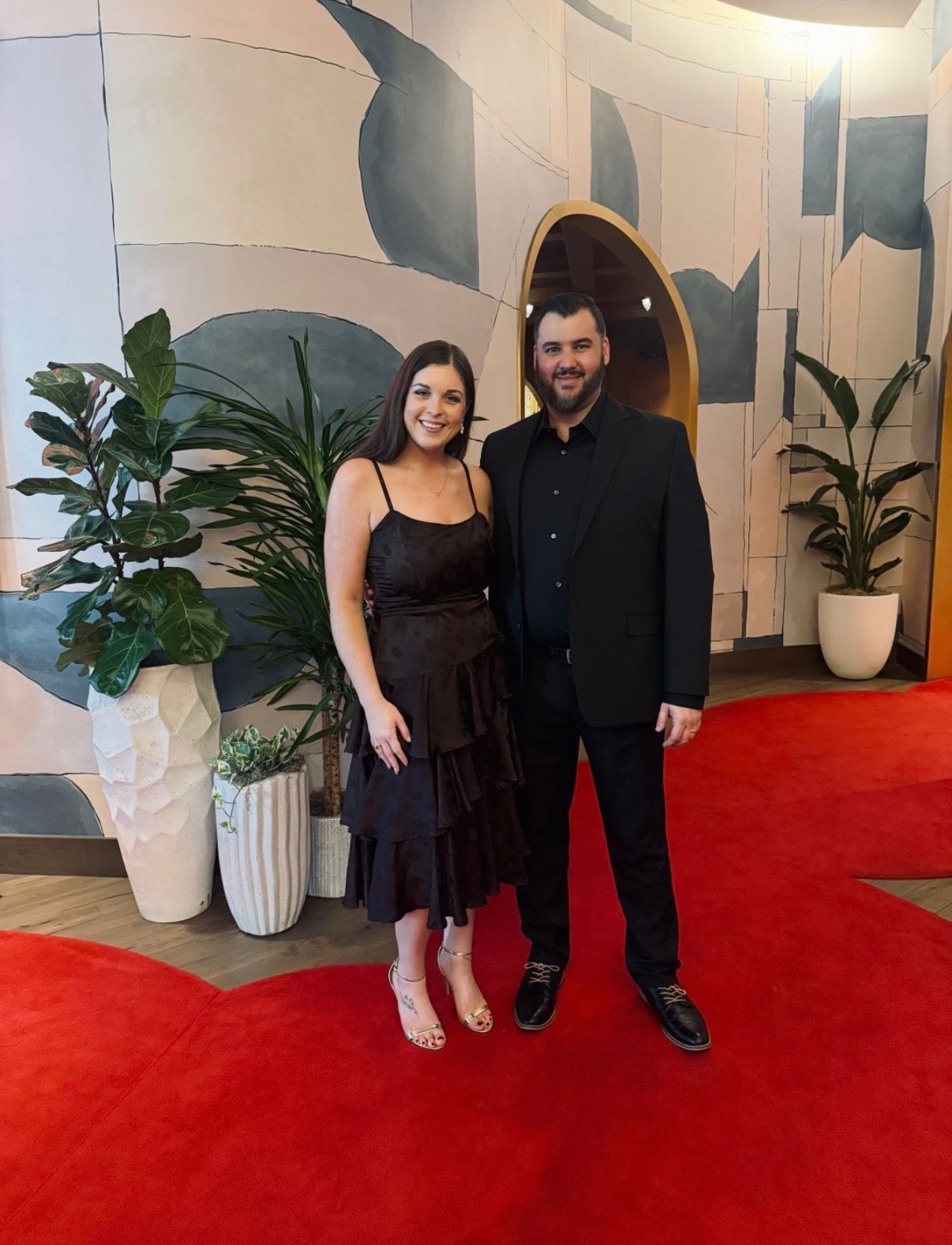 Two people posing on a red carpet in front of a patterned wall and potted plants.