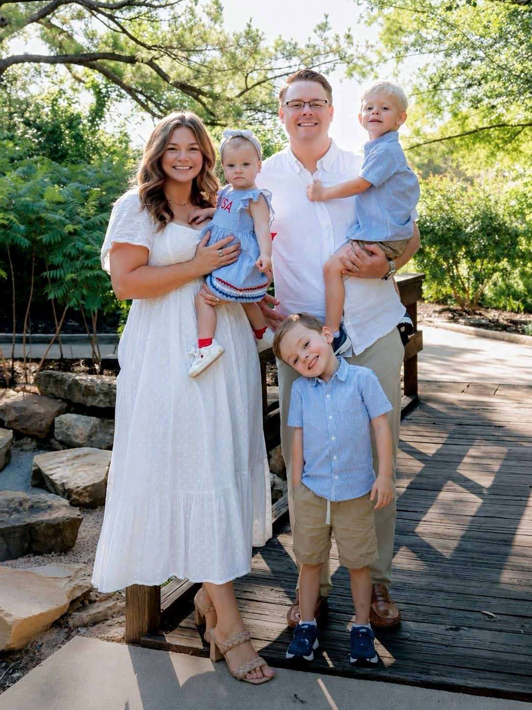Family of four smiling outdoors on a stone path, holding two young children in a sunny garden