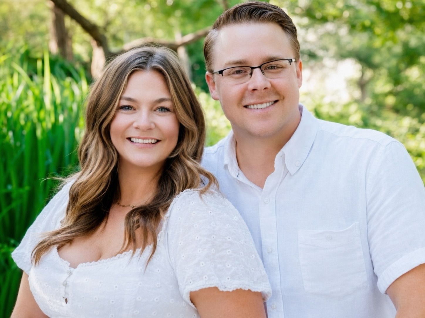 Two people smiling outdoors in white shirts, standing in a green garden background
